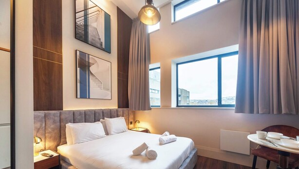 Une chambre d’hôtel avec un grand lit, un bureau avec une chaise, une fenêtre avec des rideaux et des œuvres d’art accrochées aux murs.
