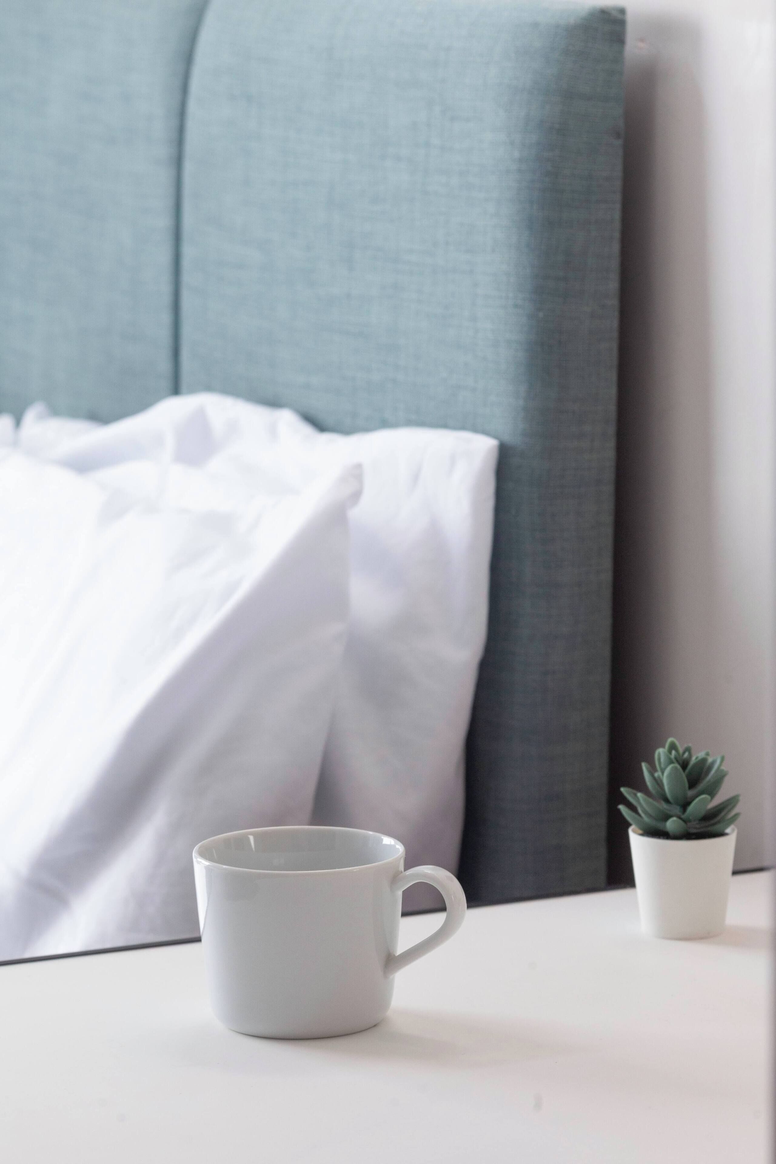 Una cama con ropa de cama blanca, un cabecero gris, una taza blanca sobre una mesa blanca y una pequeña planta en maceta.