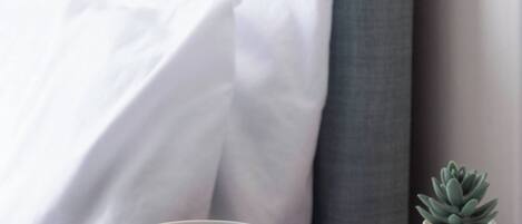 Una cama con ropa de cama blanca, un cabecero gris, una taza blanca sobre una mesa blanca y una pequeña planta en maceta.