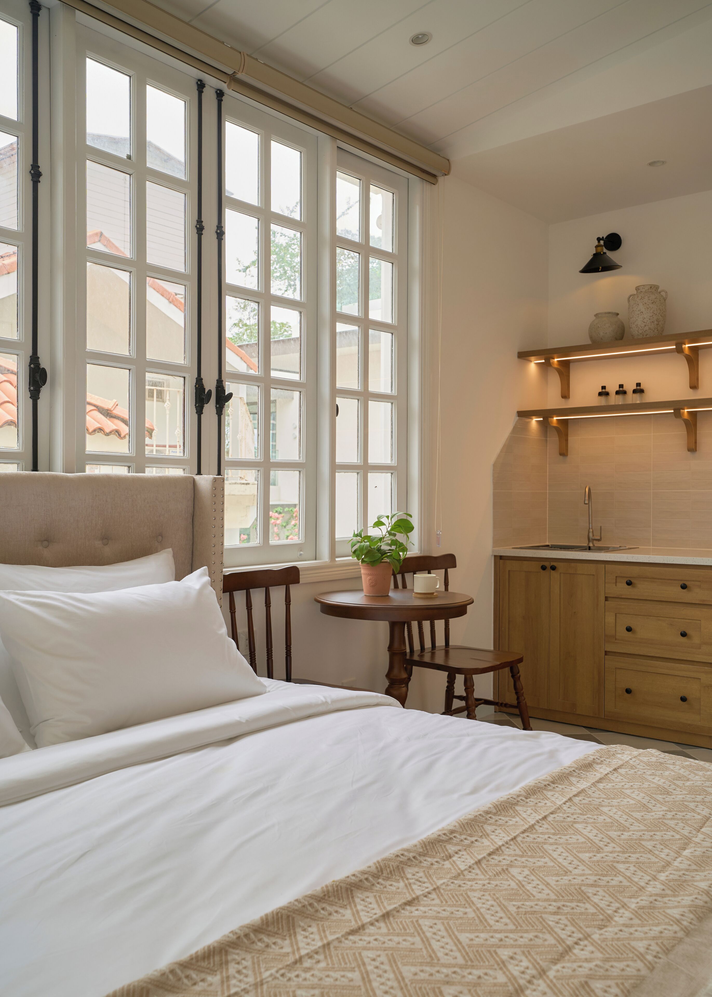 A bedroom with a bed, a bedside table with a plant, a window with blinds, and a kitchen area with cabinets and a shelf.