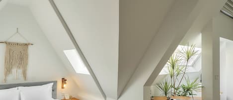A modern loft bedroom with a sloped ceiling, a bed, a seating area, and a workspace with a desk and chair.