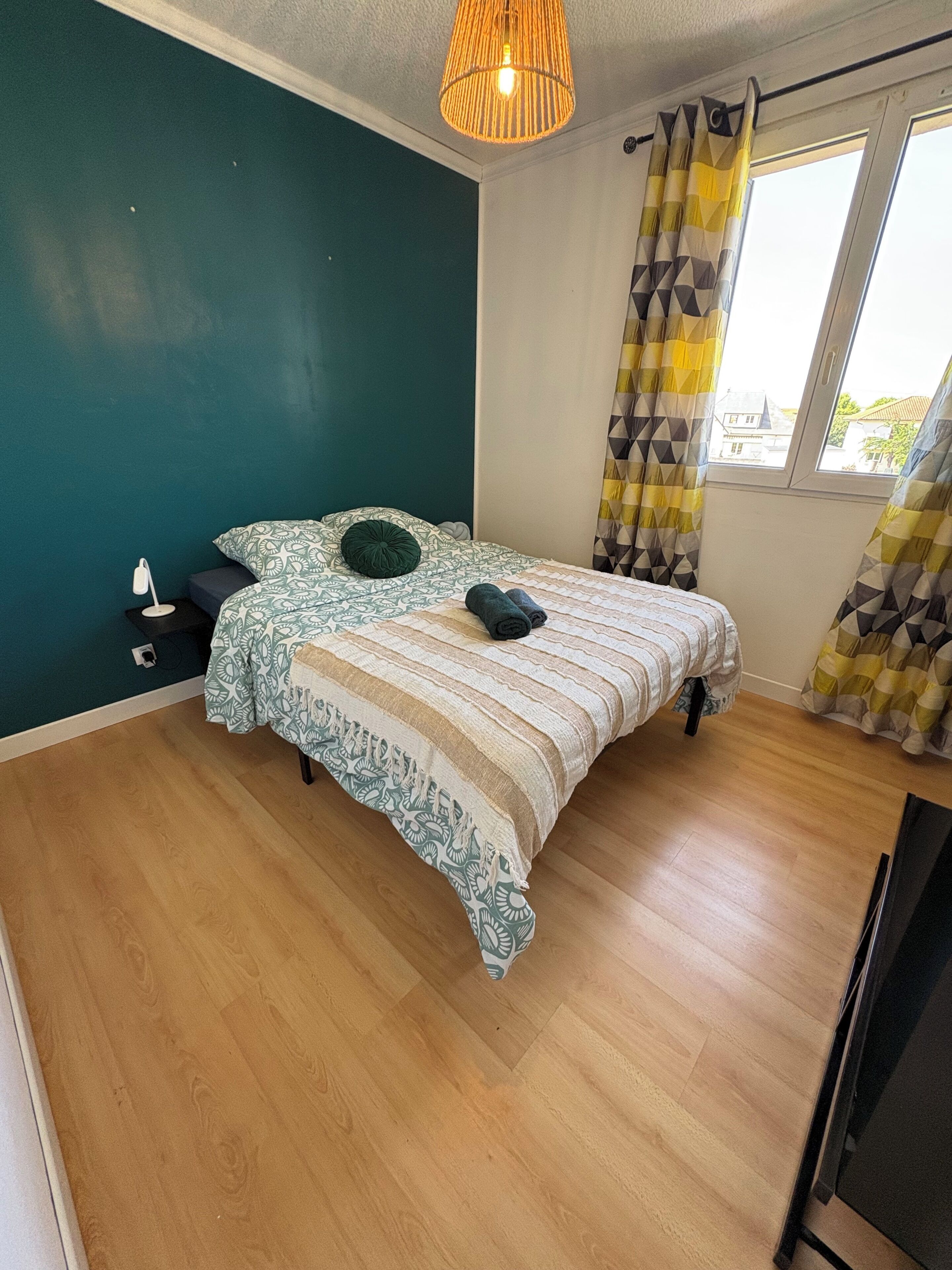 A bedroom with a bed, a bedside table, a lamp, a window with curtains, and a hanging light fixture.