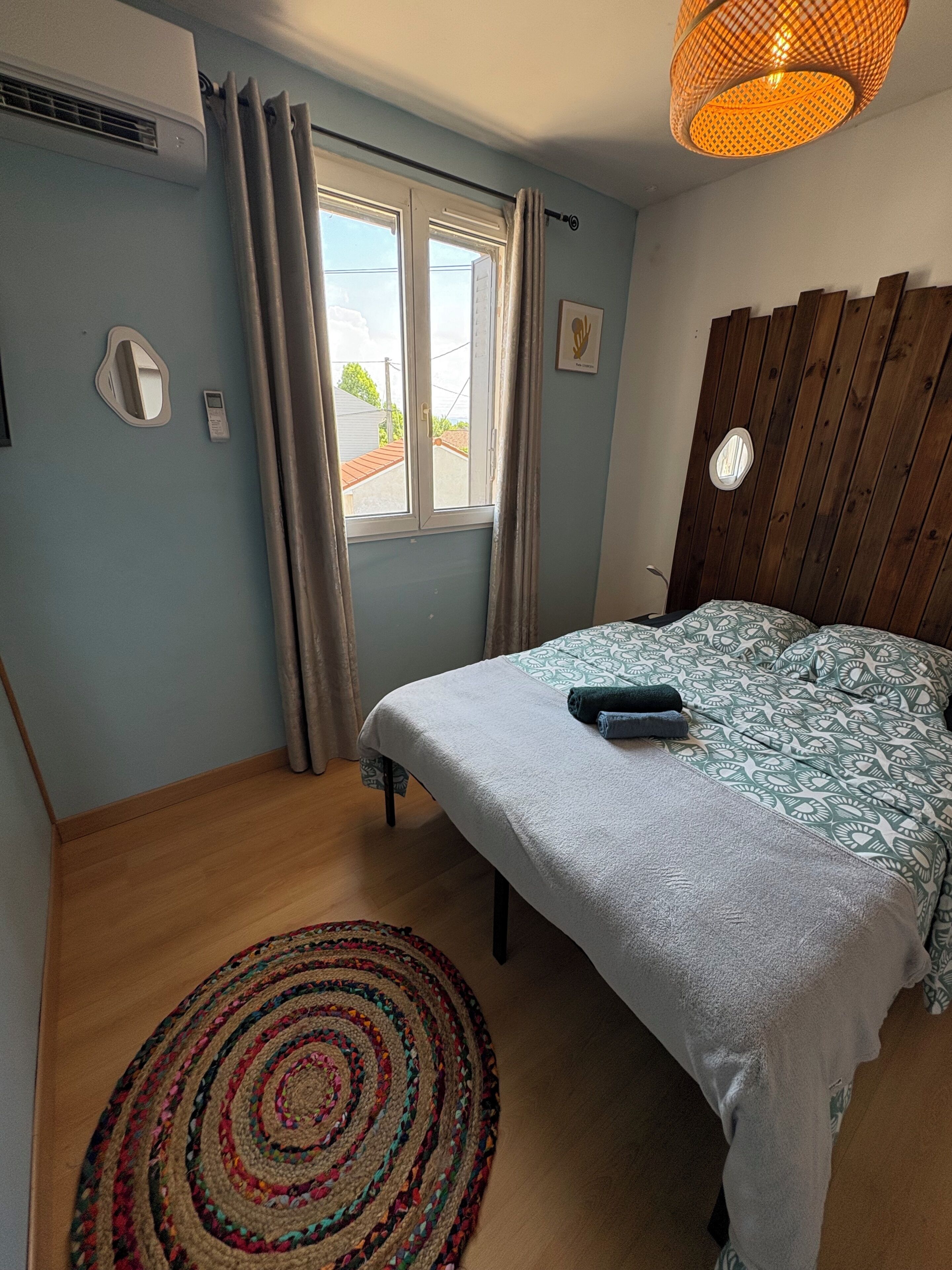 A bedroom with a bed, a window, a wooden headboard, a wall-mounted air conditioner, and a ceiling light fixture.