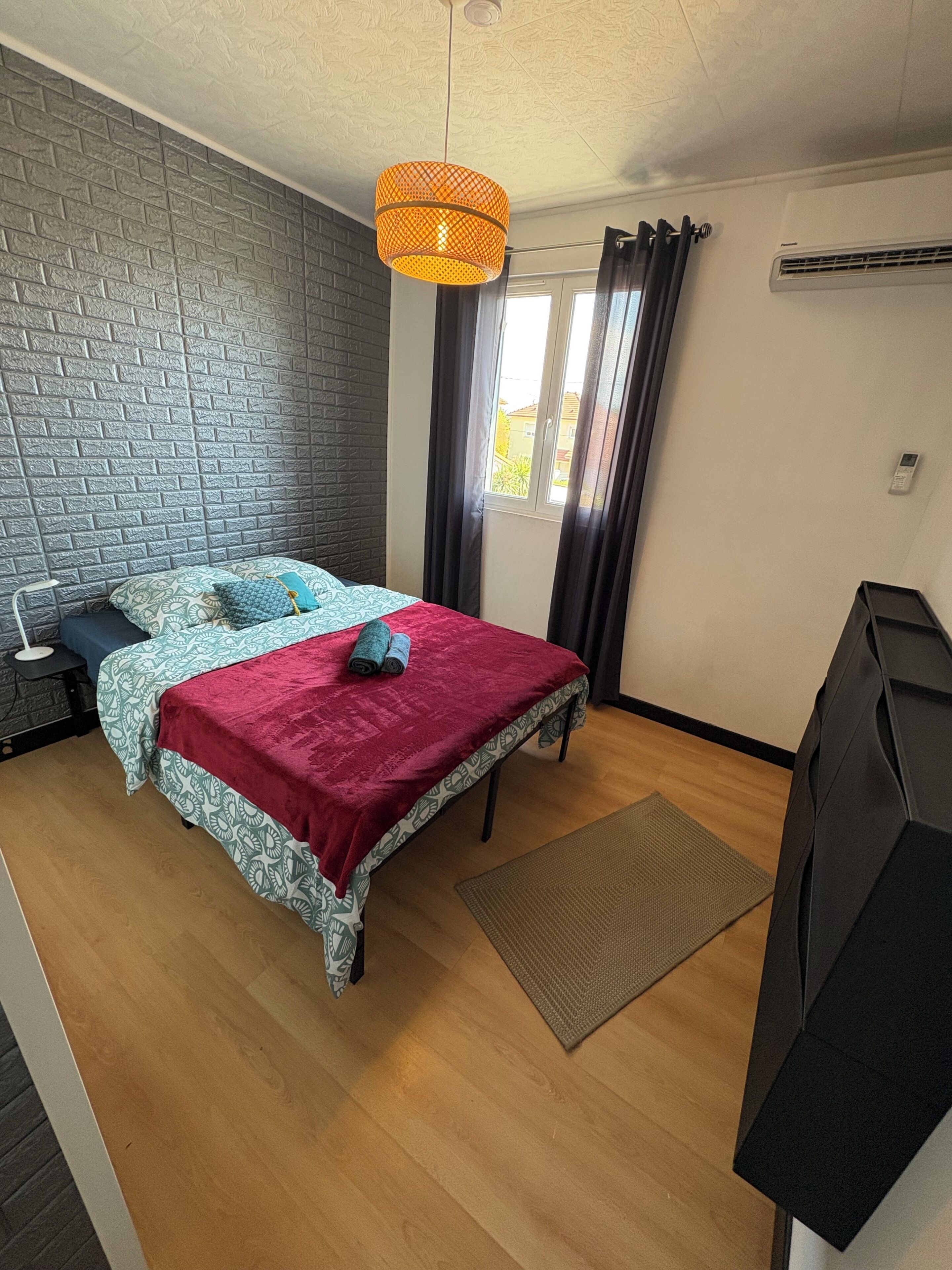 Een slaapkamer met een metselwerkmuur, een bed met beddengoed met een patroon, een rode deken, een hangende oranje lamp en een raam met gordijnen.