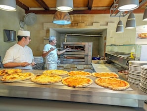 A commercial kitchen with a pizza oven, chefs, and a variety of pizzas on display. - Hotiday Torre Santo Stefano Resort (Otranto)