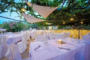 An outdoor dining area with round tables covered in white tablecloths, arranged under a canopy of trees. - Hotiday Torre Santo Stefano Resort (Otranto)