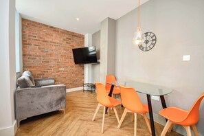 Ein modernes Wohnzimmer mit Backsteinwand, Flachbildfernseher, Glastisch und orangen Stühlen sowie einer Wanduhr.