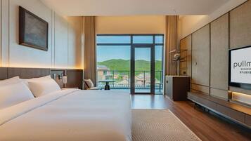 A modern hotel room with a large bed, a television, and a view of a green landscape through large windows.