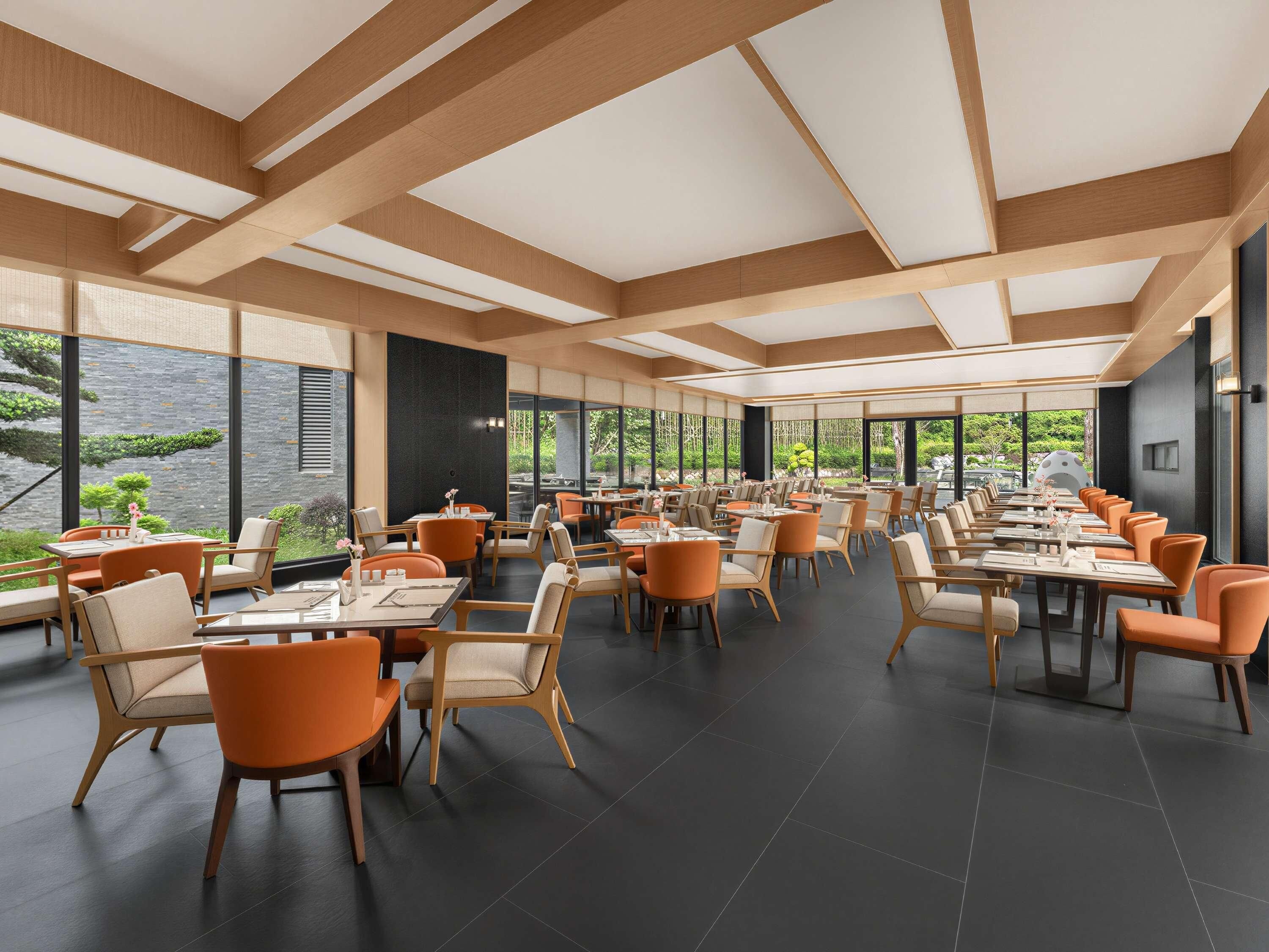 A spacious dining area with high ceilings and large windows overlooking a garden. Tables are arranged in rows with orange chairs.