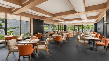 A spacious dining area with high ceilings and large windows overlooking a garden. Tables are arranged in rows with orange chairs.