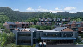 A modern hotel with a prominent glass facade, surrounded by lush greenery and hills.