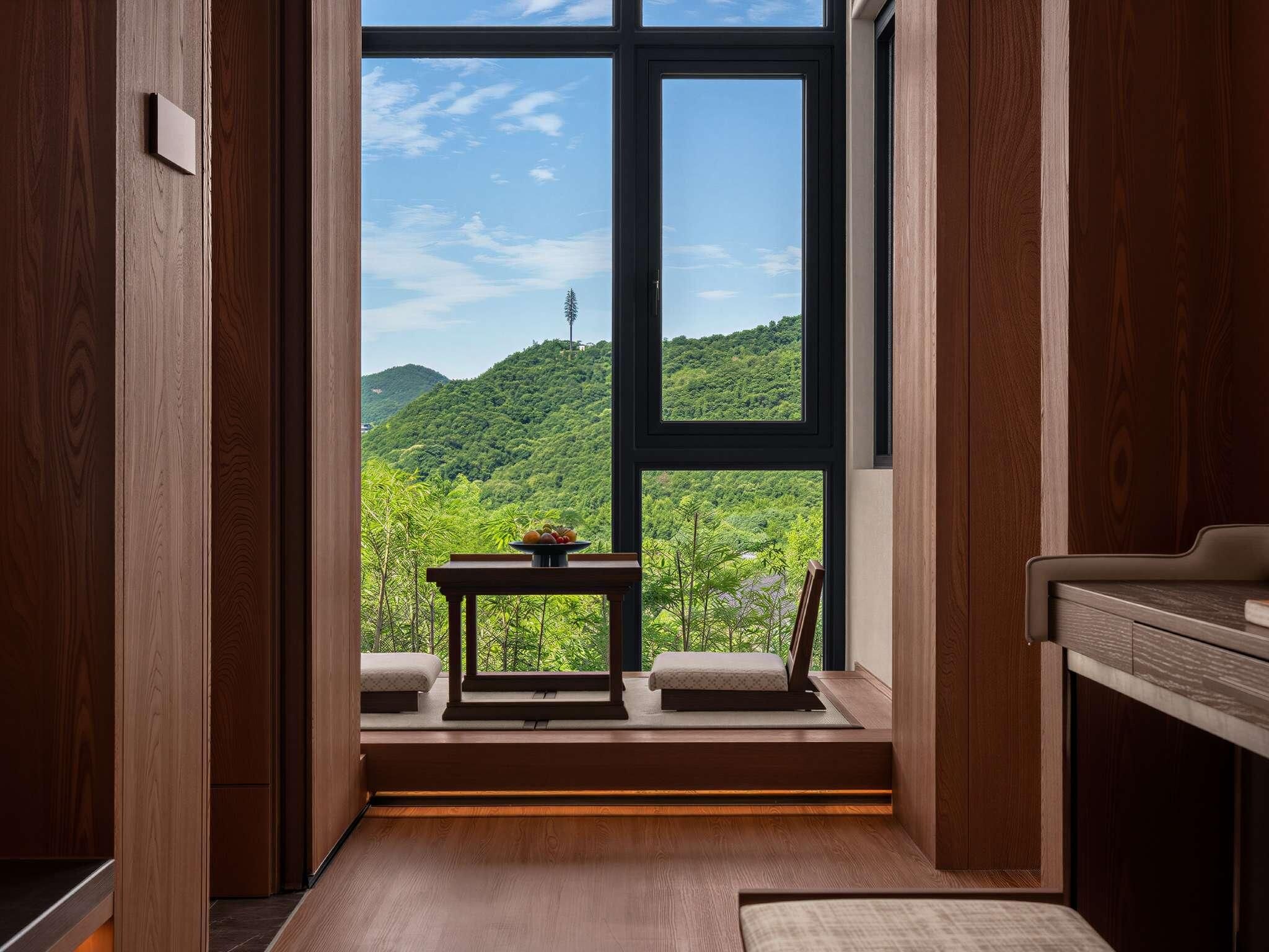 A room with a large window, wooden flooring, a small table with a fruit bowl, and a bed with a white bedspread.