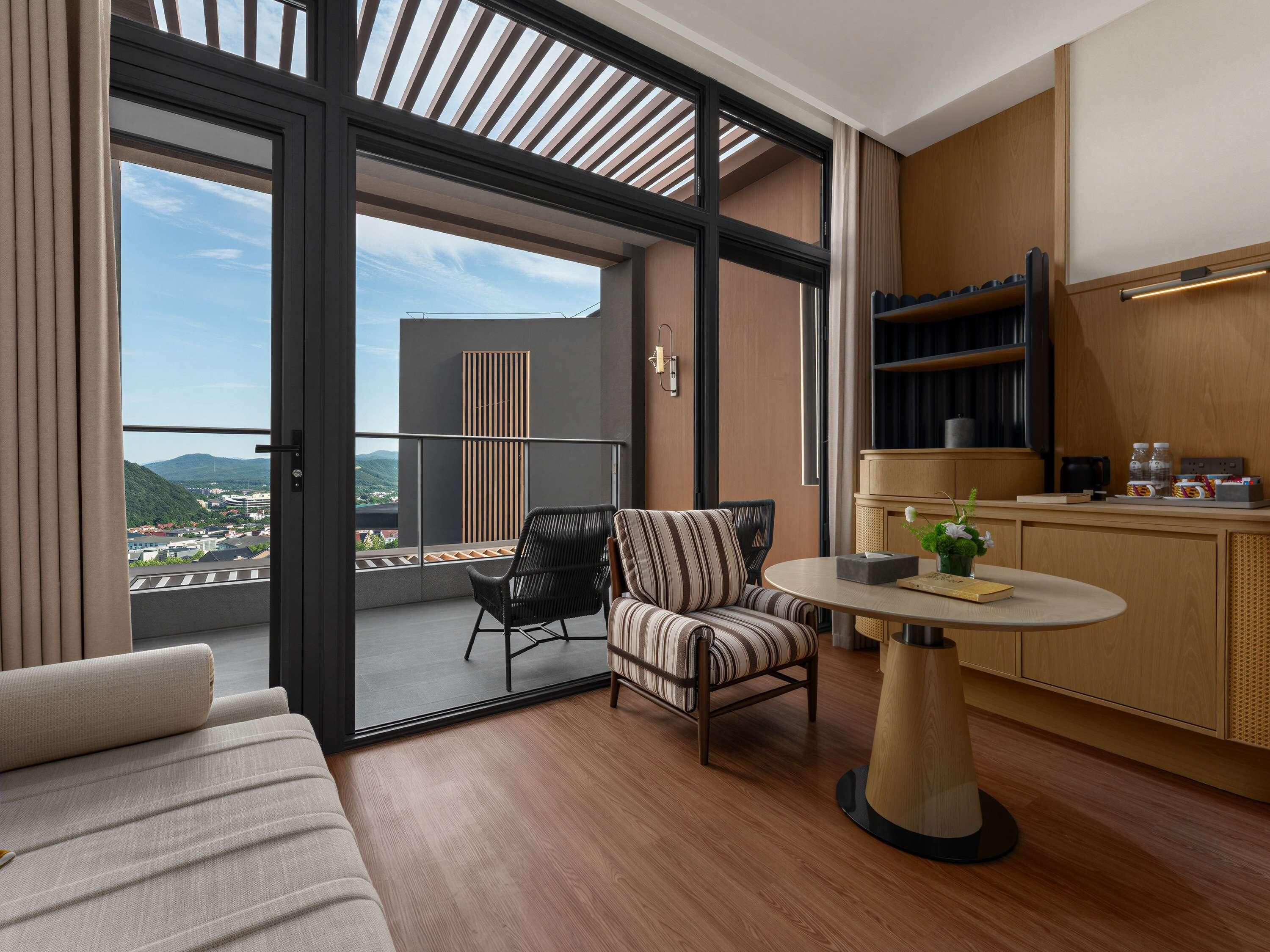 A modern hotel room with a balcony, a sofa, a chair, a small table, and a kitchenette.