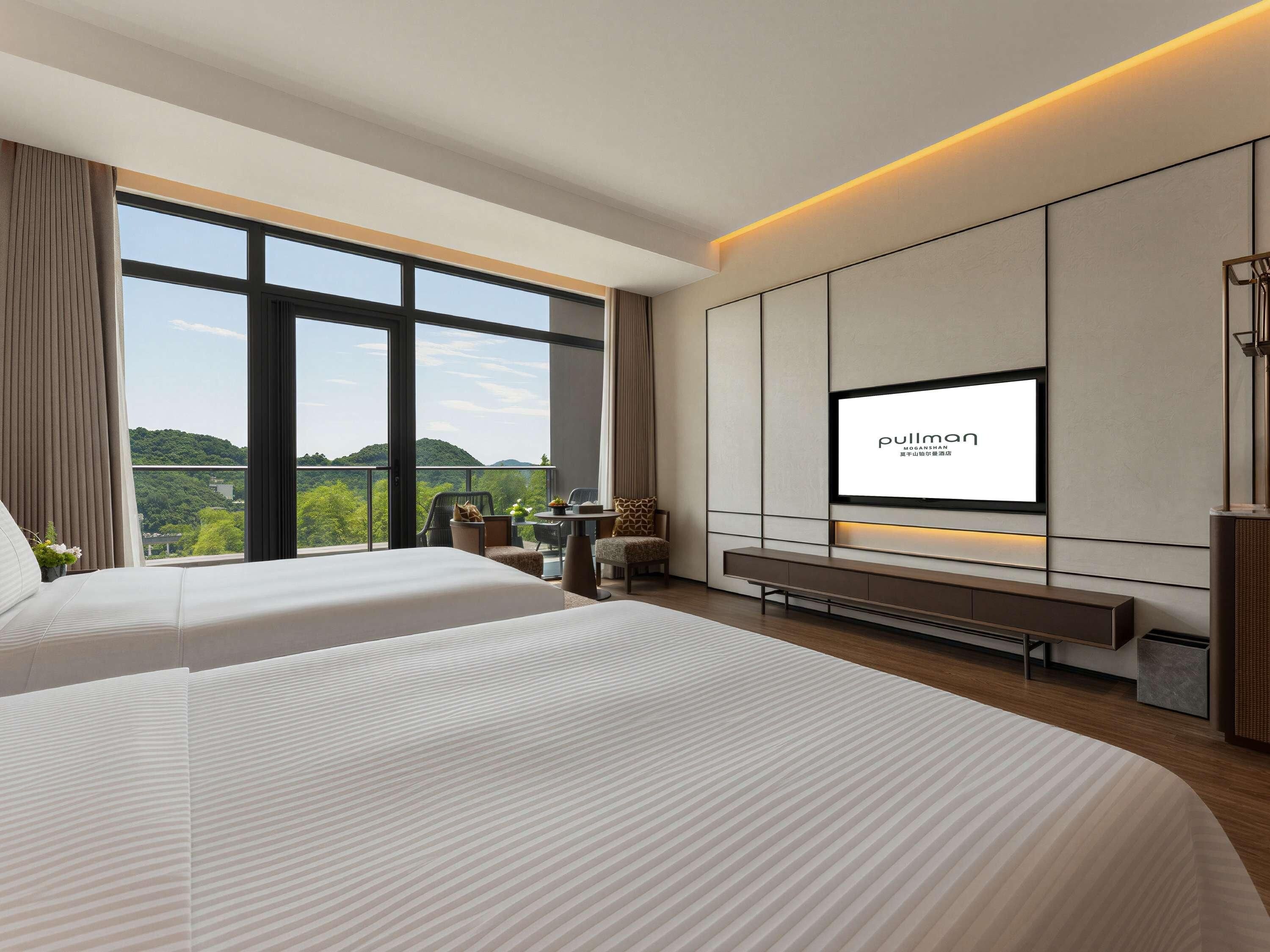 Habitación de hotel con dos camas, televisión, balcón con vistas a zonas verdes y escritorio con sillas.