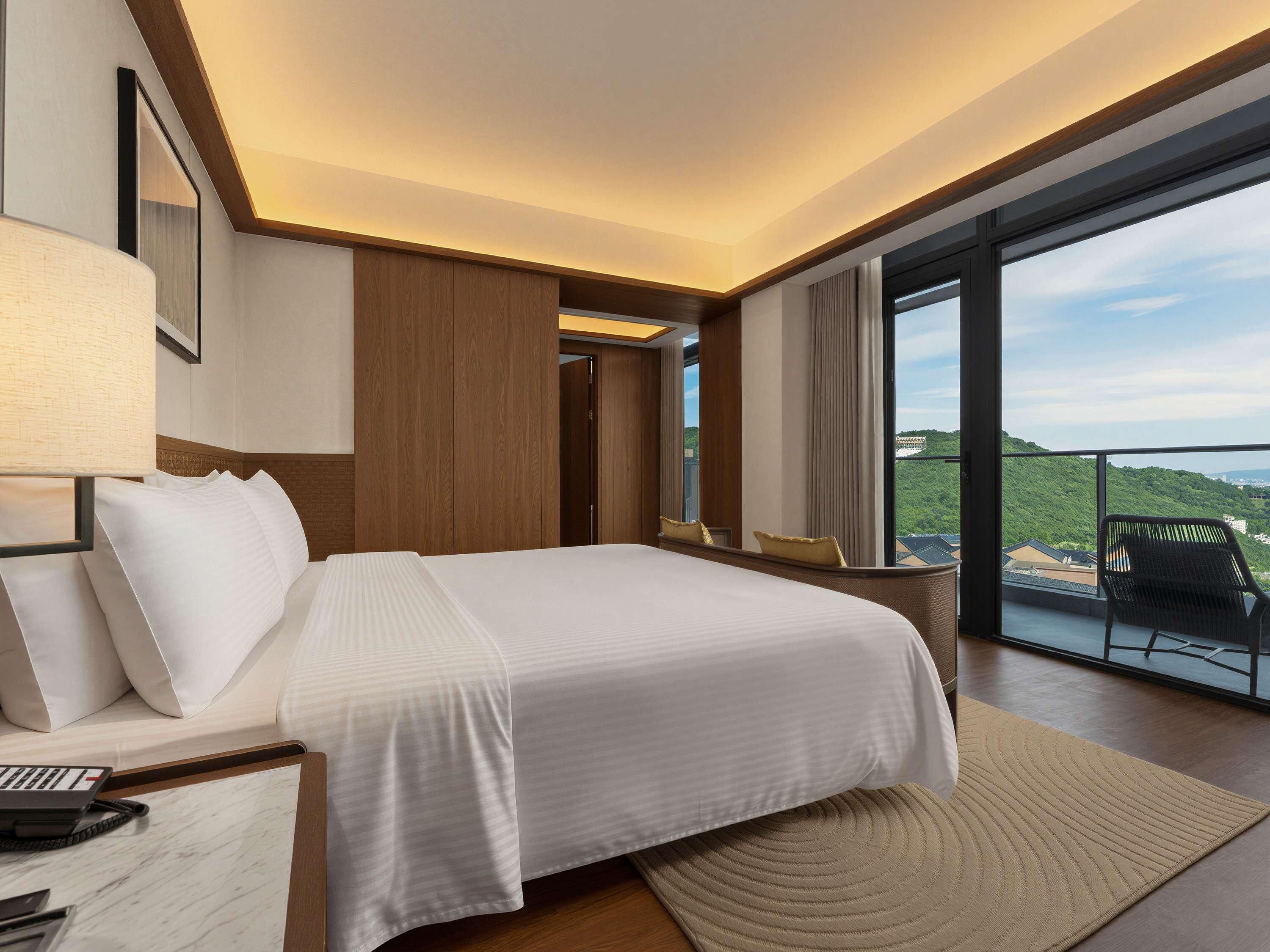Une chambre d’hôtel avec un grand lit, un bureau avec un téléphone et une vue sur un paysage verdoyant par une grande fenêtre.