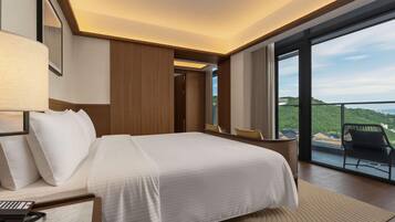 Une chambre d’hôtel avec un grand lit, un bureau avec un téléphone et une vue sur un paysage verdoyant par une grande fenêtre.