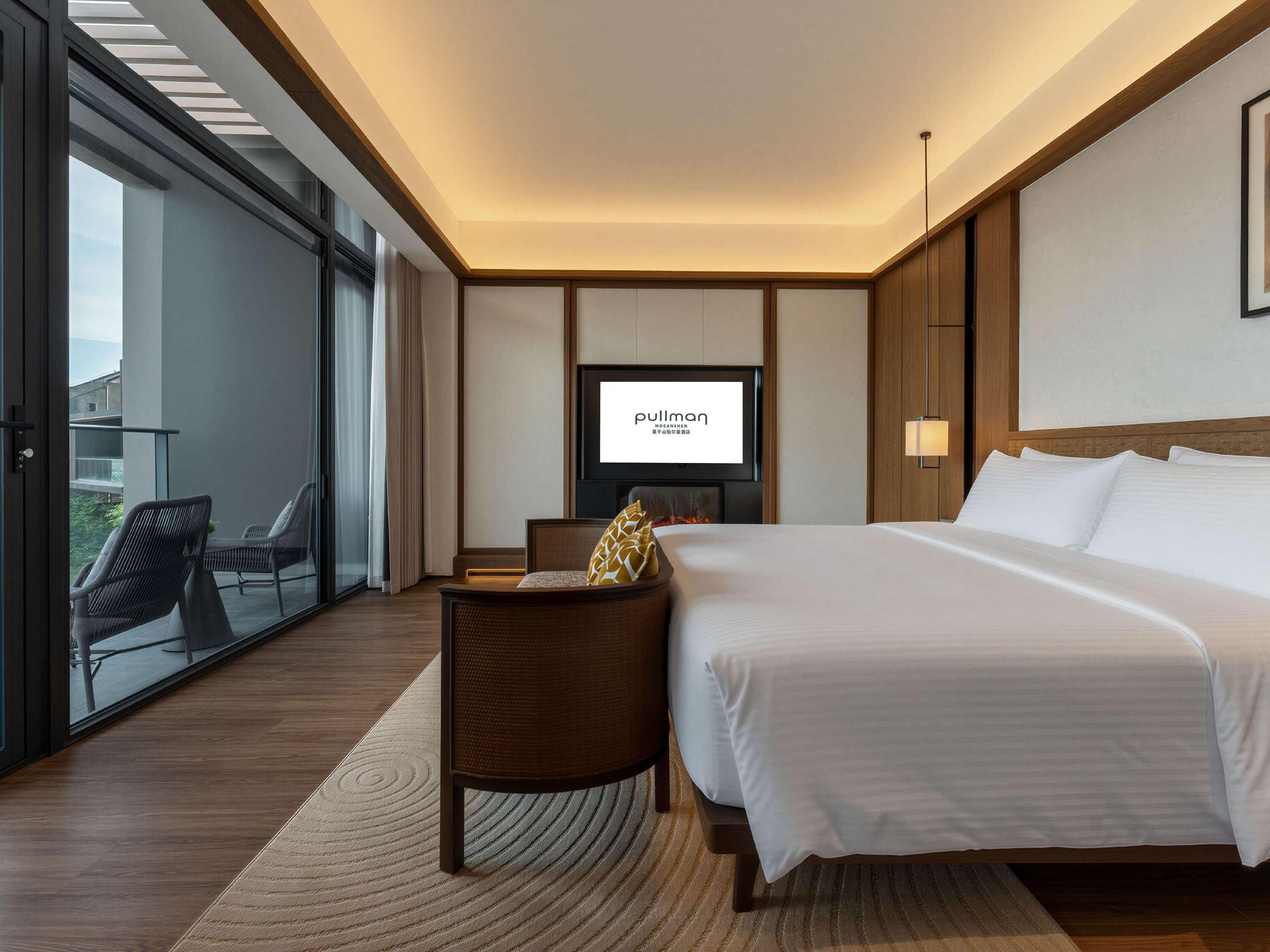 Habitación de hotel con cama, televisor, zona de estar y vistas a través de un ventanal.
