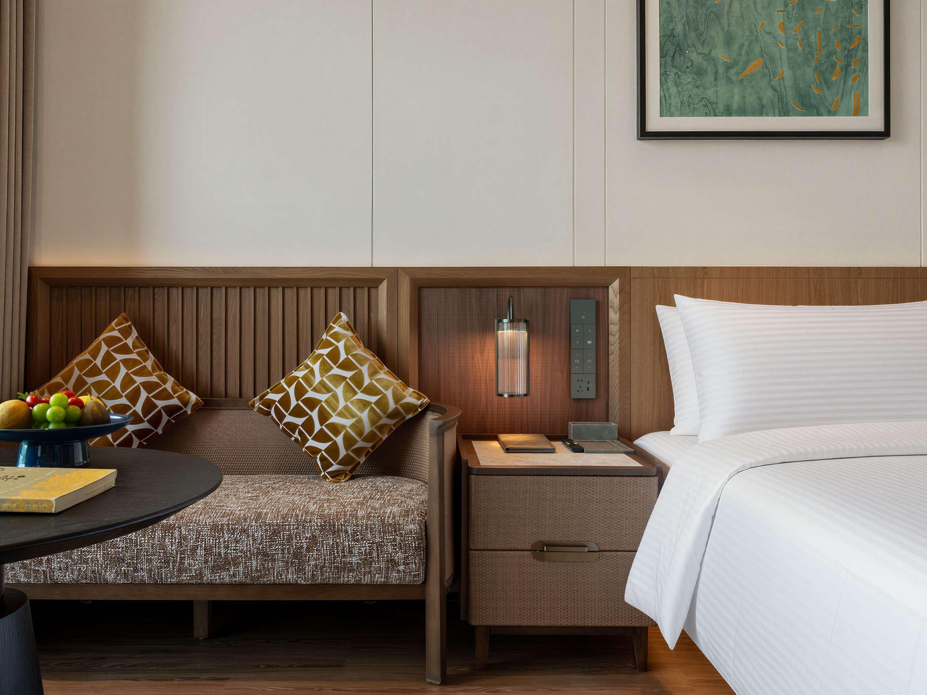 Habitación de hotel con cama, mesita de noche, lámpara, sillón y una pequeña mesa con frutero.