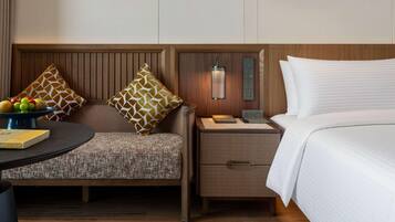 Habitación de hotel con cama, mesita de noche, lámpara, sillón y una pequeña mesa con frutero.