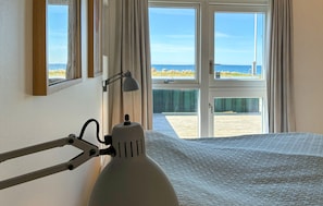 Ein Bett mit weißer Strukturdecke, eine Schreibtischlampe und ein Fenster mit Blick auf Strand und Meer.