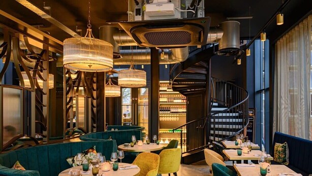 L’intérieur d’un restaurant avec des tables rondes, des sièges recouverts d’un tissu vert, des suspensions lumineuses et un escalier en colimaçon.