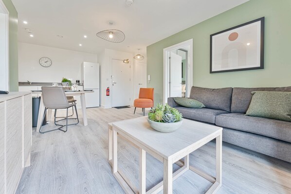 A modern living room with a grey sofa, a white coffee table with a plant, a dining area with a chair and table, and a kitchen in the background. - Stay Zeni, Hardwick House Derby Lovely 2 Bed Flat (Derby)