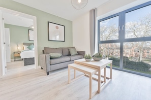 A modern living room with a sofa, a table, and a large window with a view of buildings. - Stay Zeni, Hardwick House Derby Lovely 2 Bed Flat (Derby)