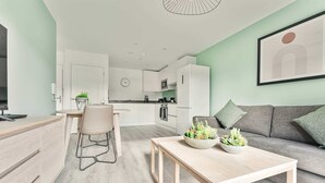 Un salon moderne avec un canapé, un coin repas avec une table et des chaises, une cuisine en arrière-plan et une horloge au mur.