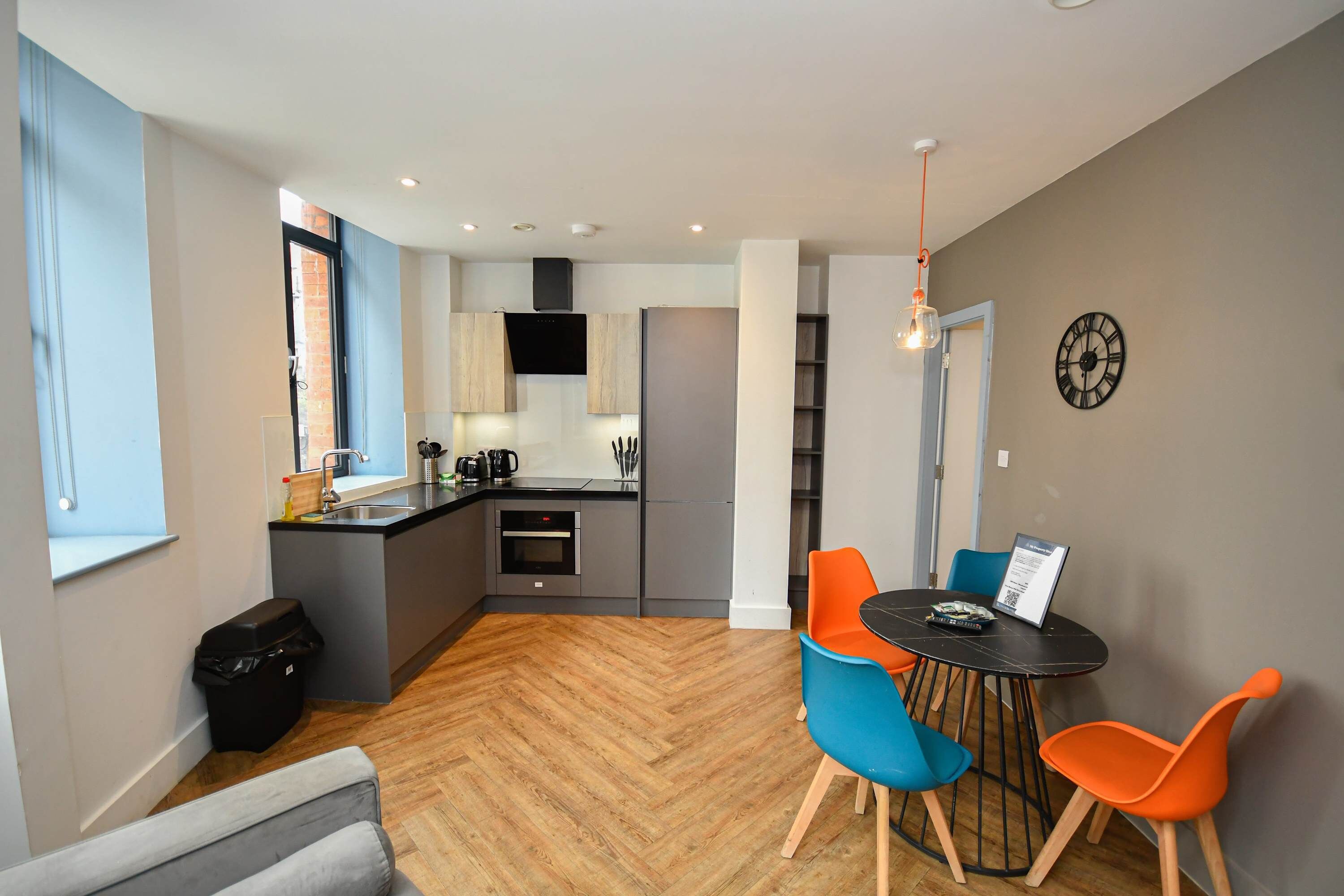 A modern kitchen with a central island, a dining area with colorful chairs, and a clock on the wall.