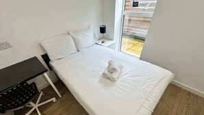 En snyggt bäddad säng med vita lakan och hoprullade handdukar, ett litet sängbord med en lampa, ett skrivbord med en stol och ett fönster med utsikt över omgivningen.