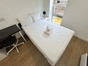 A neatly made bed with white linens and rolled towels, a small bedside table with a lamp, a desk with a chair, and a window with a view of the outside. - Budget En-suites, Central Liverpool (Liverpool)