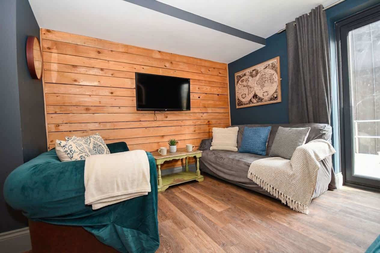 A room with a wooden wall, a flat-screen TV, a sofa, a bed, and a window with curtains.