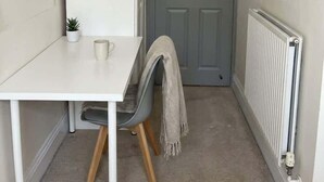 Una habitación pequeña y estrecha con una mesa blanca, una silla gris, un radiador y una puerta gris.
