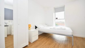 Un dormitori minimalista amb un llit individual, una tauleta de nit, una làmpada i una finestra que dona vistes a edificis.