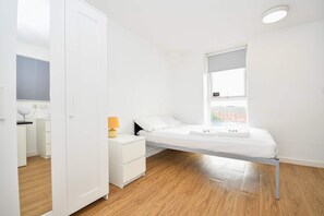 Ein schlichter Schlafzimmer mit einem Einzelbett, einem Nachttisch, einer Lampe und einem Fenster mit Blick auf Gebäude.