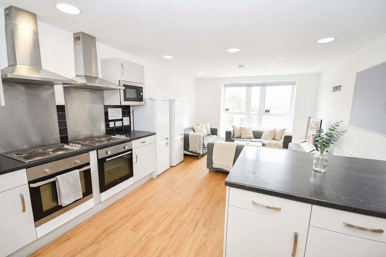 A modern kitchen with stainless steel appliances, a dining area with a black countertop, and a view of a living room with a sofa and a window.