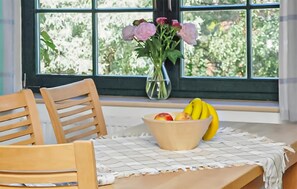 Ein Holzhocker, ein Tisch mit Unterlage, eine Obstschale und eine Vase mit Blumen an einem Fenster.