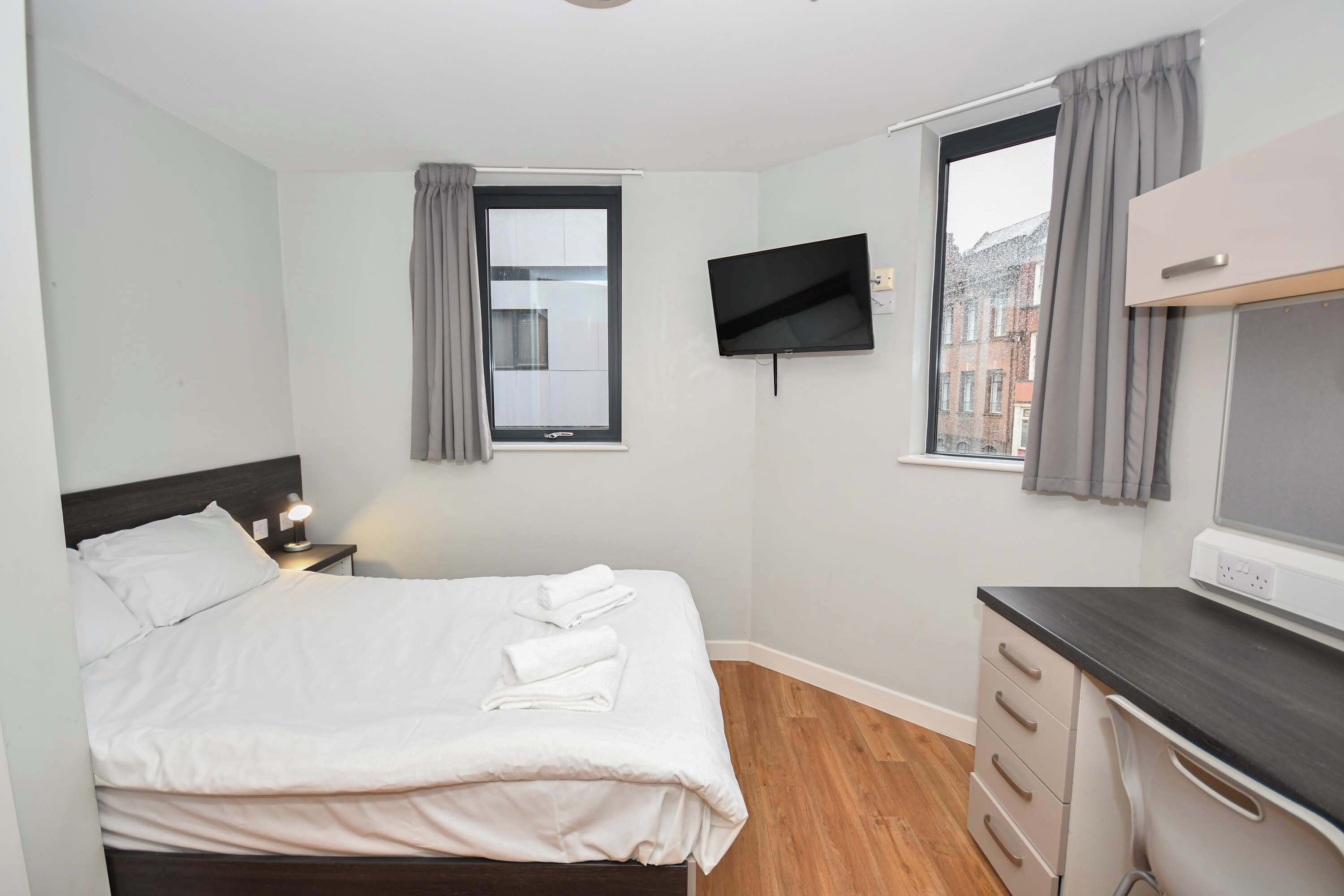 A hotel room with a bed, a desk, a TV, a window with curtains, and a view of buildings.