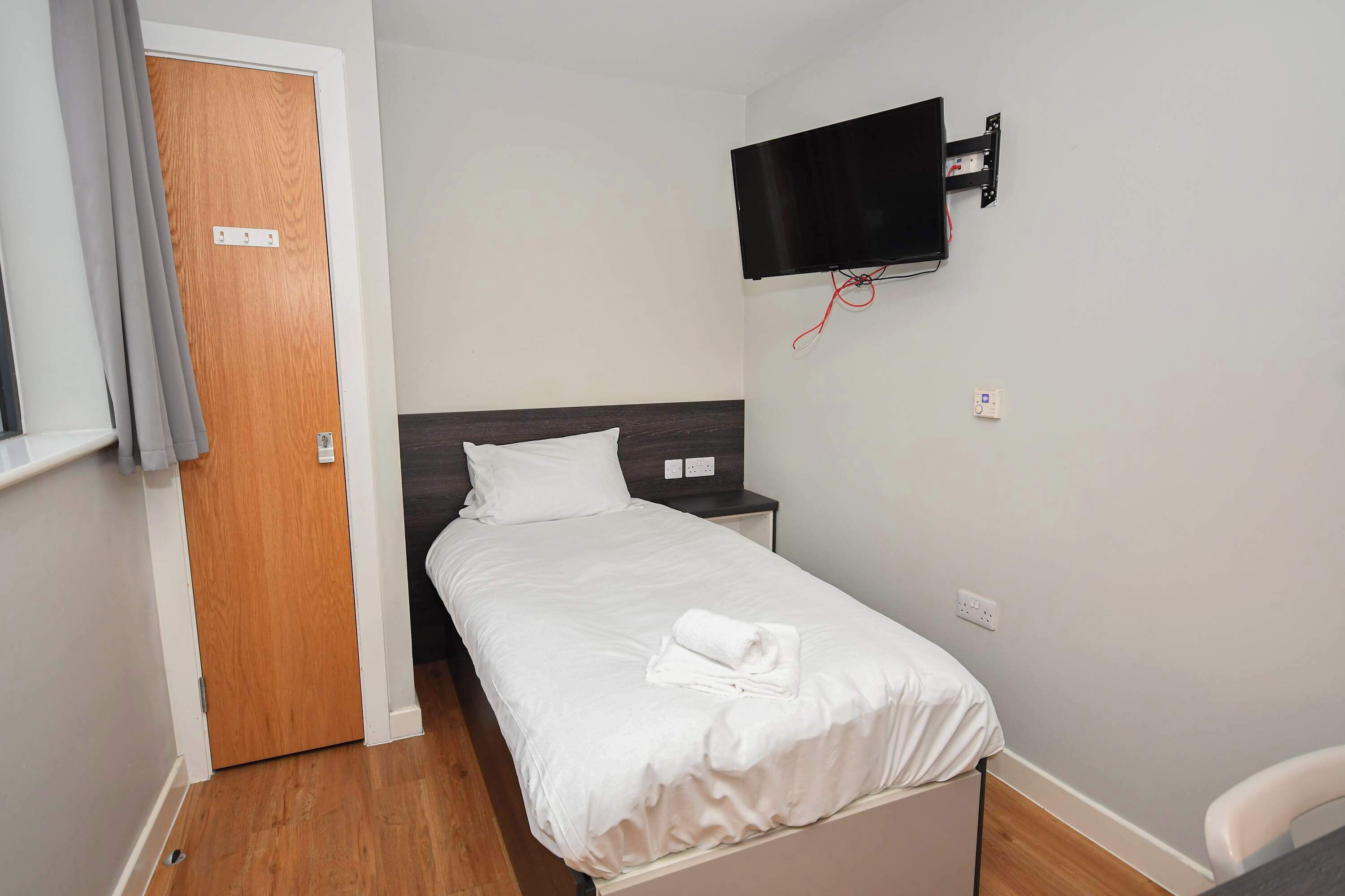 A neatly made bed with white linens, a wooden door, a mounted TV, and a small bedside table.