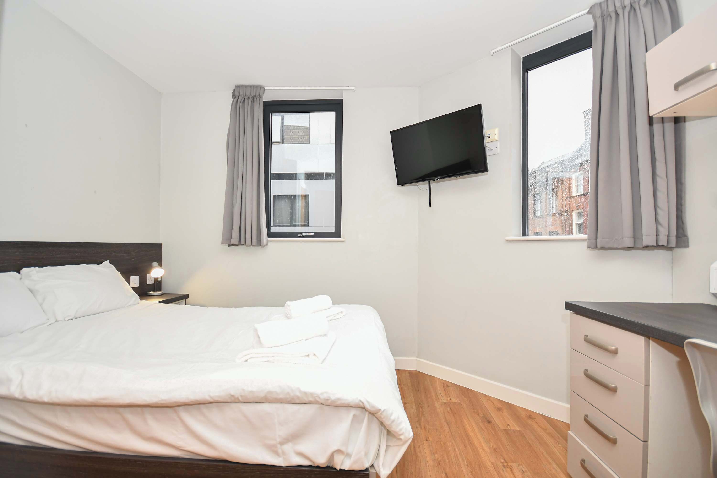 A bedroom with a bed, a TV mounted on the wall, a desk, and a window with curtains.