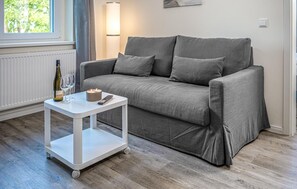 A grey sofa with cushions, a white side table with a remote control, a candle, and a bottle of wine on the floor. - 1 bedroom amazing apartment in Barth (Barth)