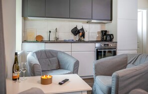 Eine moderne Küche mit grauen Schränken, einer Kaffeemaschine und einer Mikrowelle. Zwei graue Sessel und ein Tisch mit Kerze und Fernbedienung.