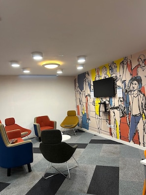 A room with a modern design, featuring a flat-screen TV mounted on the wall, a variety of colorful chairs, and a patterned mural.