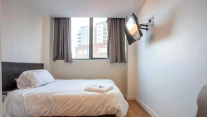 A bedroom with a bed, a TV mounted on the wall, a window with curtains, and a view of buildings outside.