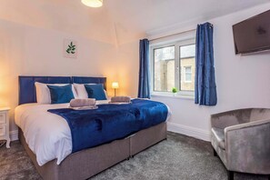 A bedroom with a bed, bedside table, lamp, curtains, and a TV.