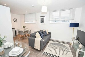 A modern living room with a grey sofa, dining table, and a large window with blinds.