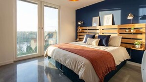 A bedroom with a bed, a wooden headboard, a bedside table with a lamp, and a view through the window. - Modern & Spacious 2BED Apartment by Old Trafford (Manchester)