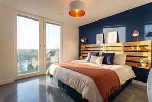 A bedroom with a bed, a wooden headboard, a bedside table with a lamp, and a view through the window. - Modern & Spacious 2BED Apartment by Old Trafford (Manchester)