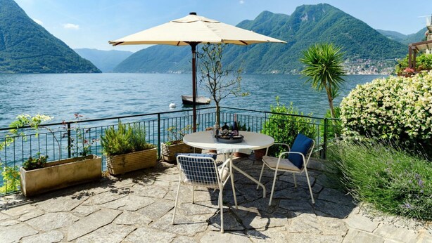 Eine Terrasse mit Tisch und Stühlen, Sonnenschirm und Blick auf See und Berge.