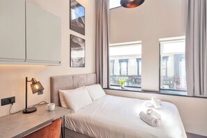 A hotel room with a bed, a bedside table with a lamp, a window with a view of buildings, and a wall-mounted artwork. - Central Hastings Studio Apartment Near Seafront (East Sussex)